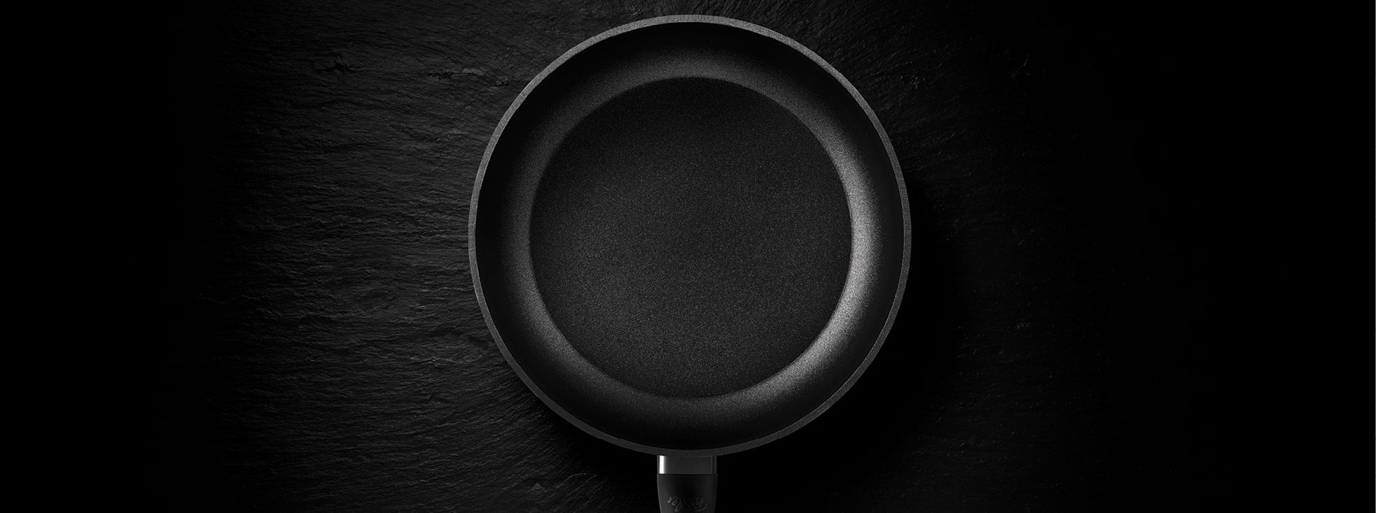Eine schwarze antihaftbeschichtete Bratpfanne von Fissler, von oben gesehen, auf einer dunkel strukturierten Oberfläche - präzise Technik trifft auf zeitloses Design.