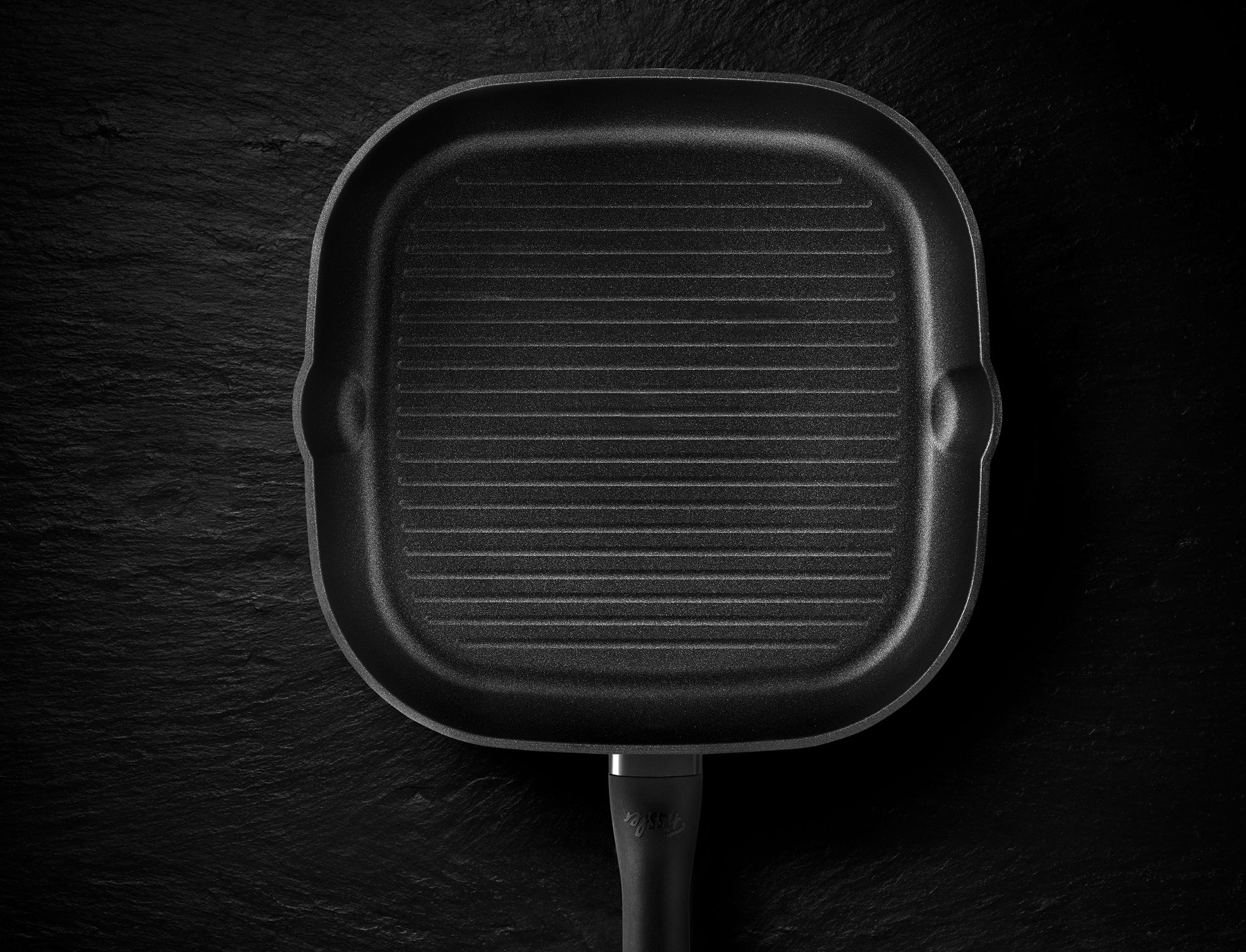 Schwarze quadratische Grillpfanne von Fissler mit Rillen, Präzisionsfertigung in Deutschland, abgebildet von oben mit Griff und zwei Ausgießern.