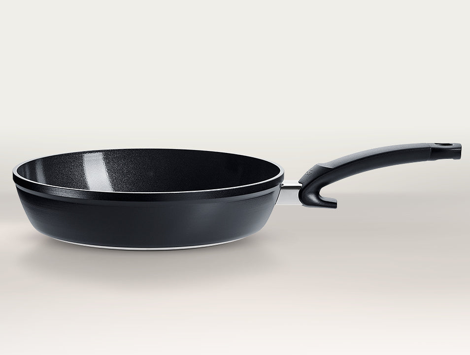 Schwarze antihaftbeschichtete Bratpfanne von Fissler mit dem charakteristischen gebogenen Griff, hergestellt in Deutschland, vor einem hellgrauen Hintergrund.