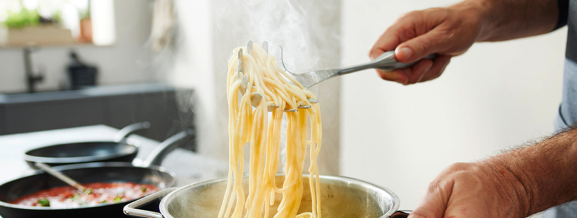 Eine Person hebt Spaghetti mit einer Zange aus einem Fissler-Edelstahltopf, im Hintergrund Kochtöpfe, die deutsches Präzisionskochgeschirr zeigen.