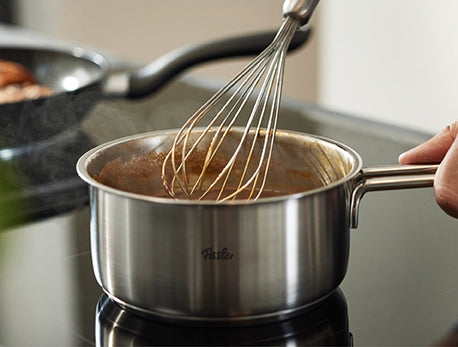 Eine Hand rührt die Soße mit einem Schneebesen in einem Fissler-Edelstahltopf auf dem Herd, Dampf steigt auf; ein weiterer Fissler-Topf steht dahinter.
