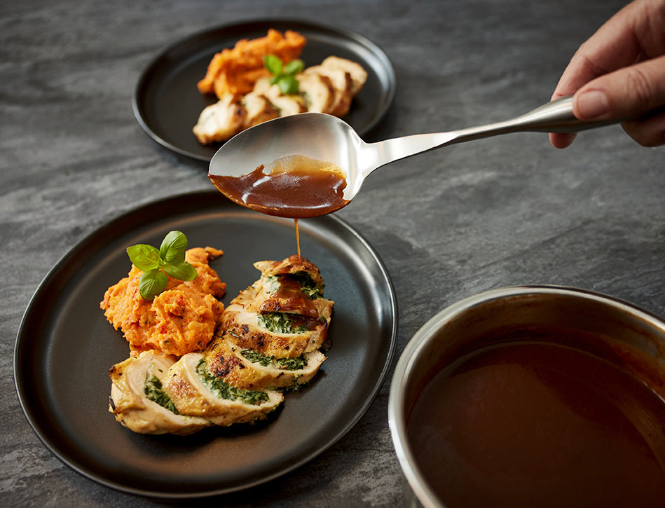 Eine Hand träufelt Sauce auf Hähnchen und Süßkartoffeln auf feinem Porzellan, das mit Präzision in Fissler-Kochgeschirr hergestellt wird.