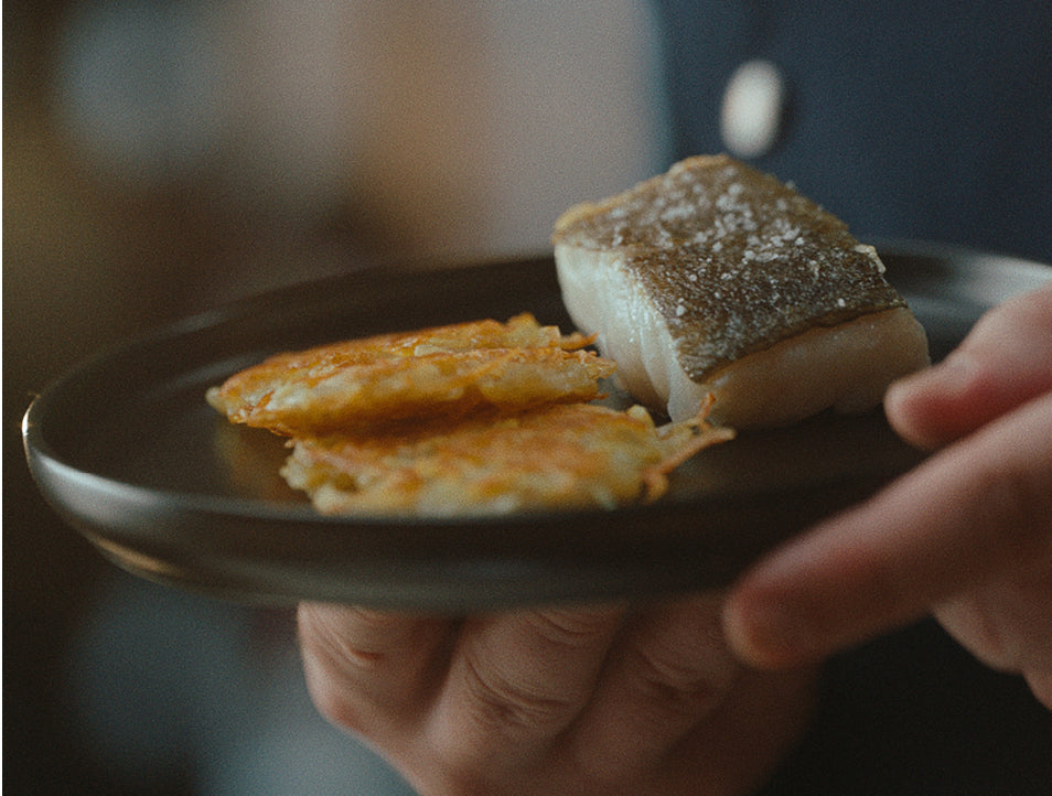Eine Person präsentiert einen dunklen Teller mit goldenen, knusprigen Kartoffelpuffern und perfekt gebratenem Fisch - zubereitet in hochwertigem Fissler-Kochgeschirr.