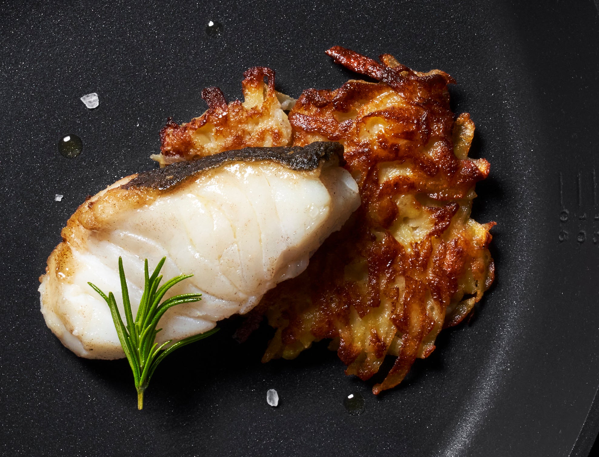 Fachmännisch gebratener weißer Fisch und goldene Kartoffelrösti auf einem schwarzen Teller, zubereitet in hochpräzisen Fissler-Kochgeschirren.