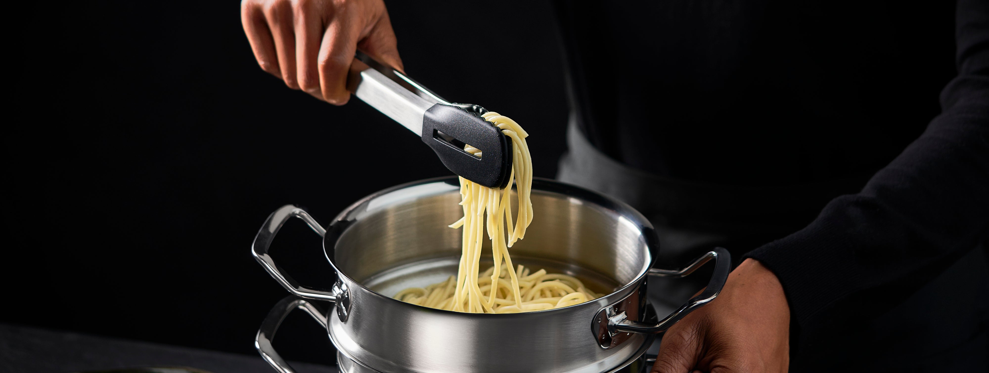 Eine Person hebt Spaghetti mit einer Zange aus einem Fissler-Edelstahltopf und präsentiert damit hochwertiges deutsches Kochgeschirr vor einem dunklen Hintergrund.
