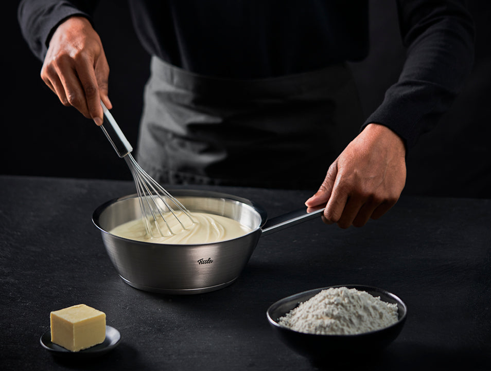 Eine Person verwendet einen Fissler-Edelstahltopf, um eine cremige Mischung zu schlagen, daneben stehen Mehl und Butter auf einer dunklen Arbeitsplatte.