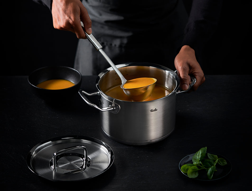 Eine Person serviert Orangensuppe aus einem Fissler-Edelstahltopf mit Deckel und frischem Basilikum auf einer dunklen Oberfläche - Präzision in jedem Detail.