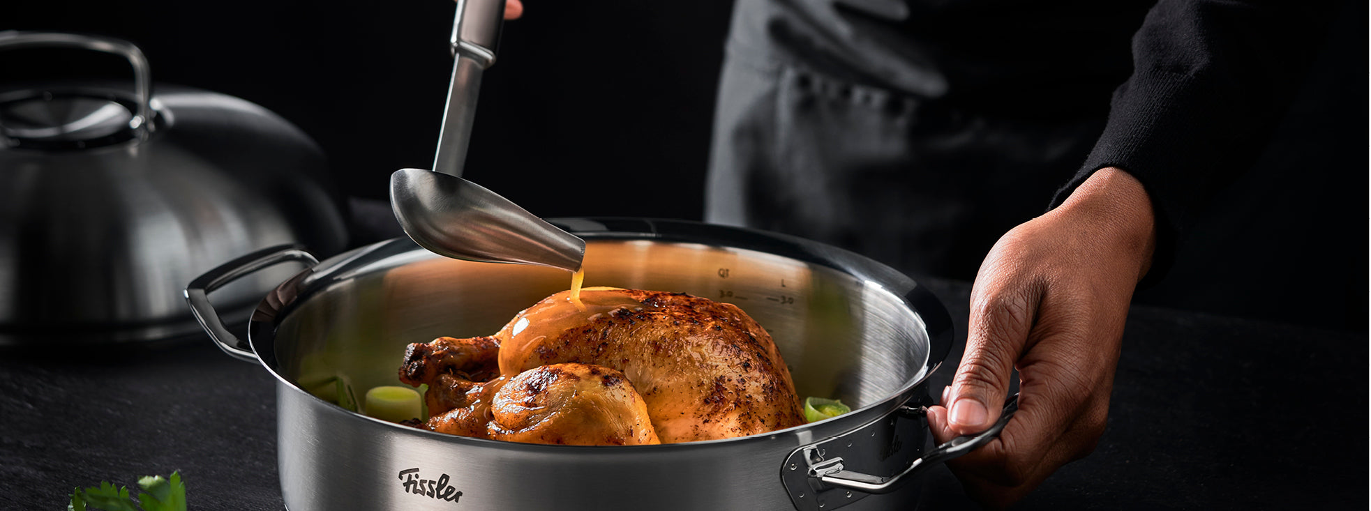 Eine Person gießt die Sauce über das gebratene Hähnchen in einem Fissler-Edelstahltopf, darunter liegt grünes Gemüse - Präzision trifft Geschmack.