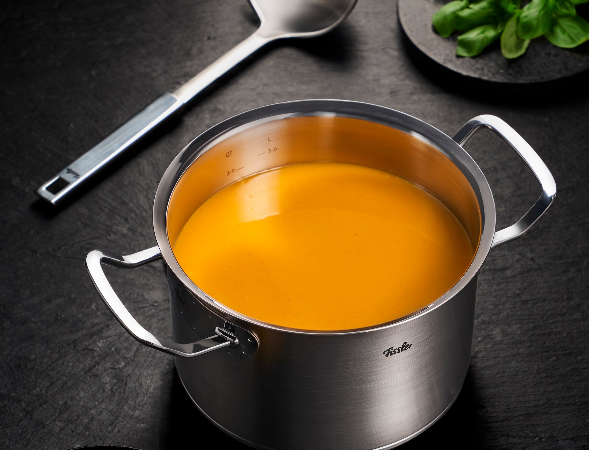 Fissler Edelstahltopf mit leuchtend orangefarbener Suppe, gepaart mit einer Präzisionskelle und frischem Basilikum für zeitlose Kücheninnovation.