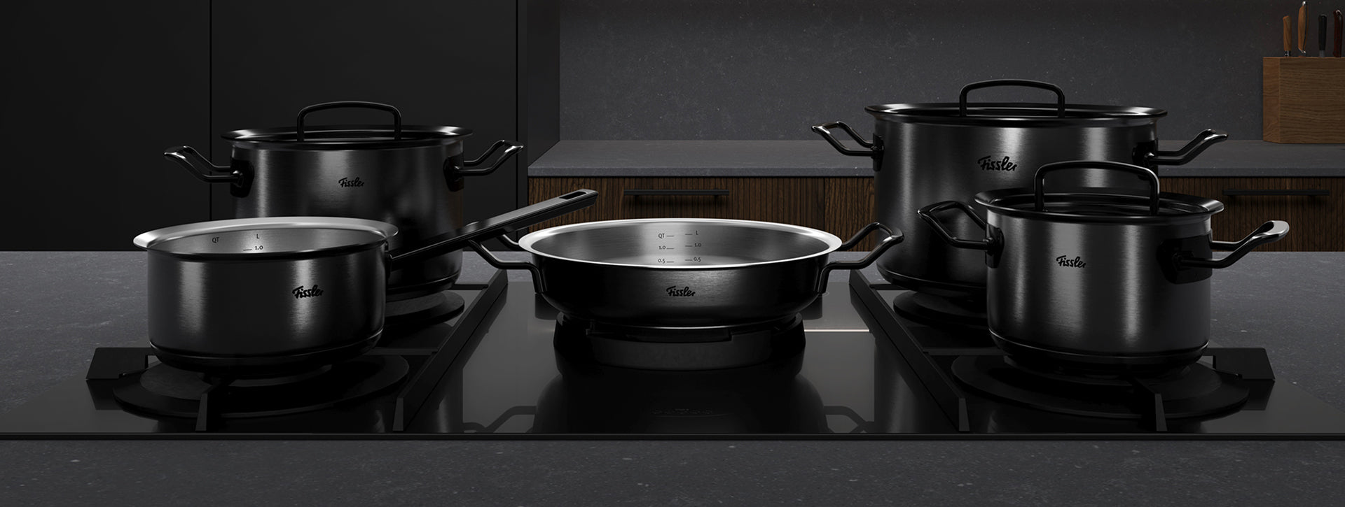 Fünf schwarze Töpfe und Pfannen mit Deckeln von Fissler auf eleganten Herdplatten in einer modernen Küche, die zeitlose deutsche Präzision und Qualität zeigen.