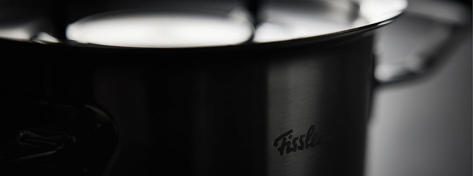 Fissler-Edelstahltopf mit reflektierendem Deckel und Logo, langlebiges Kochgeschirr aus deutscher Produktion, Griff rechts, vor dunklem Hintergrund.