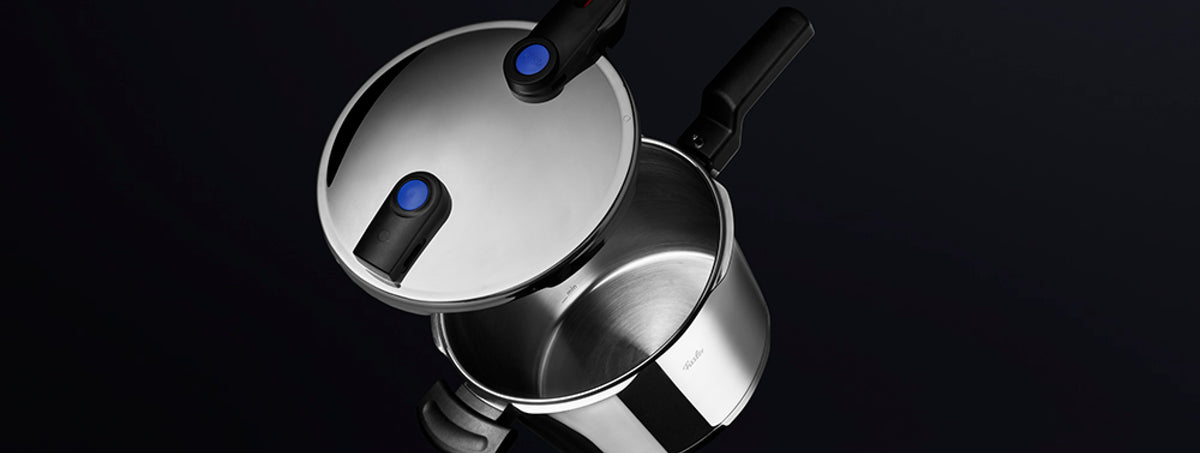 Ein Fissler-Schnellkochtopf aus Edelstahl mit schwarzen Griffen und blauen Sicherheitsknöpfen, teilweise geöffnet, ist ein Beispiel für deutsche Qualität.