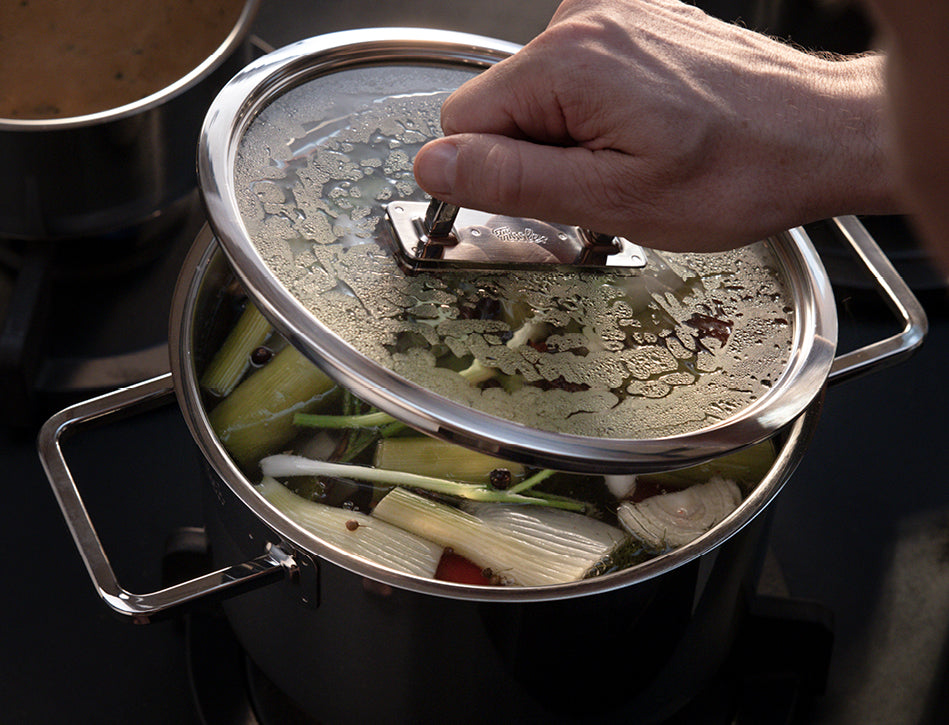 Eine Hand hebt den Deckel eines Fissler-Edelstahltopfs an und gibt den Blick frei auf dampfendes Gemüse und Kräuter, die auf dem Herd köcheln.