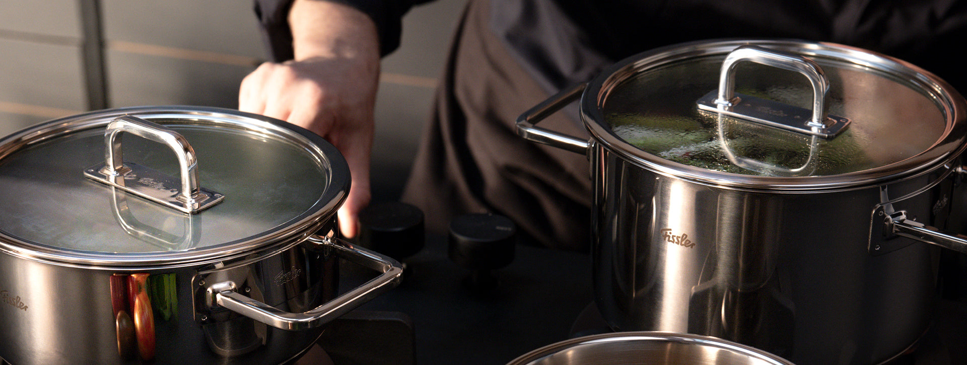 Ein Koch am Herd kümmert sich um zwei Fissler-Edelstahltöpfe mit Glasdeckeln, die in Deutschland präzisionsgefertigt wurden, während im Inneren Dampf aufsteigt.