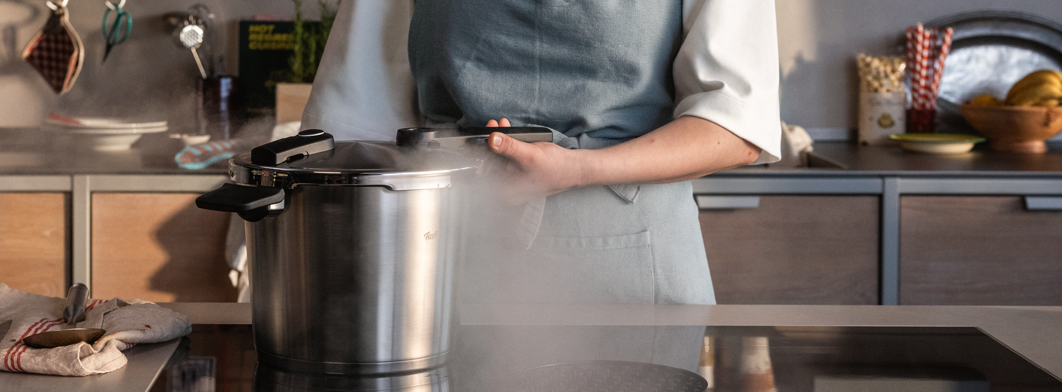 Eine Person in einer Schürze benutzt einen Fissler-Schnellkochtopf auf dem Herd; Dampf steigt auf, während deutsche Qualität auf zeitloses Design trifft.