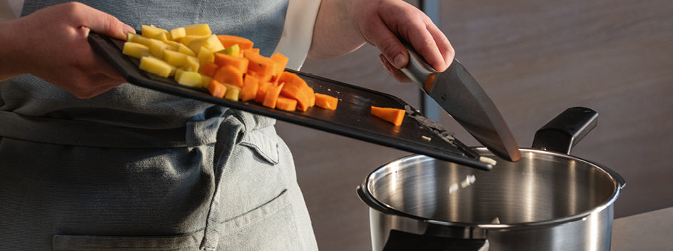 Eine Köchin in Schürze gibt frisches Gemüse in einen Fissler-Edelstahltopf, der ein Beispiel für langlebige deutsche Technik und Design ist.