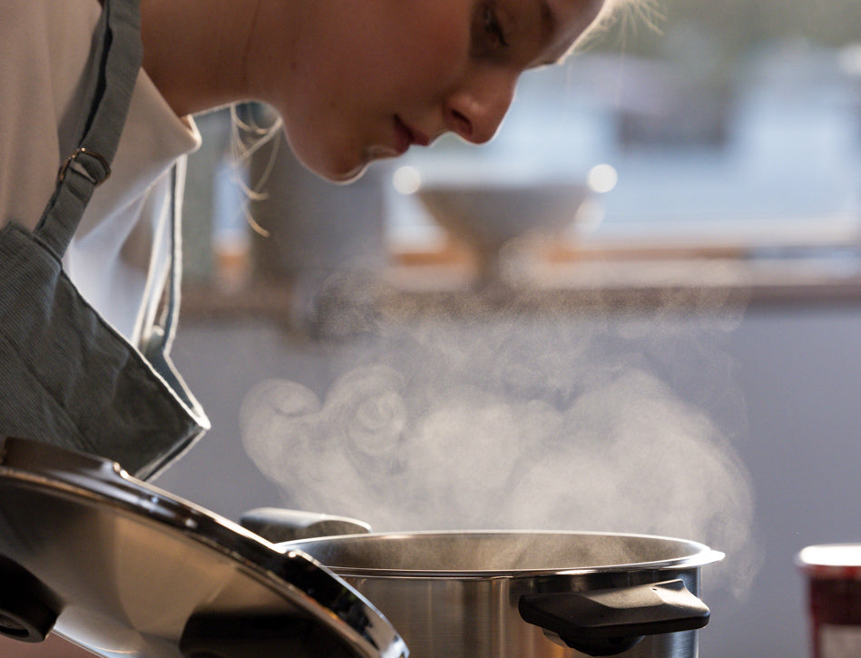 Eine Köchin in einer Schürze hebt den Deckel eines Fissler-Edelstahltopfes und genießt den aromatischen Dampf in einer hellen, modernen Küche.