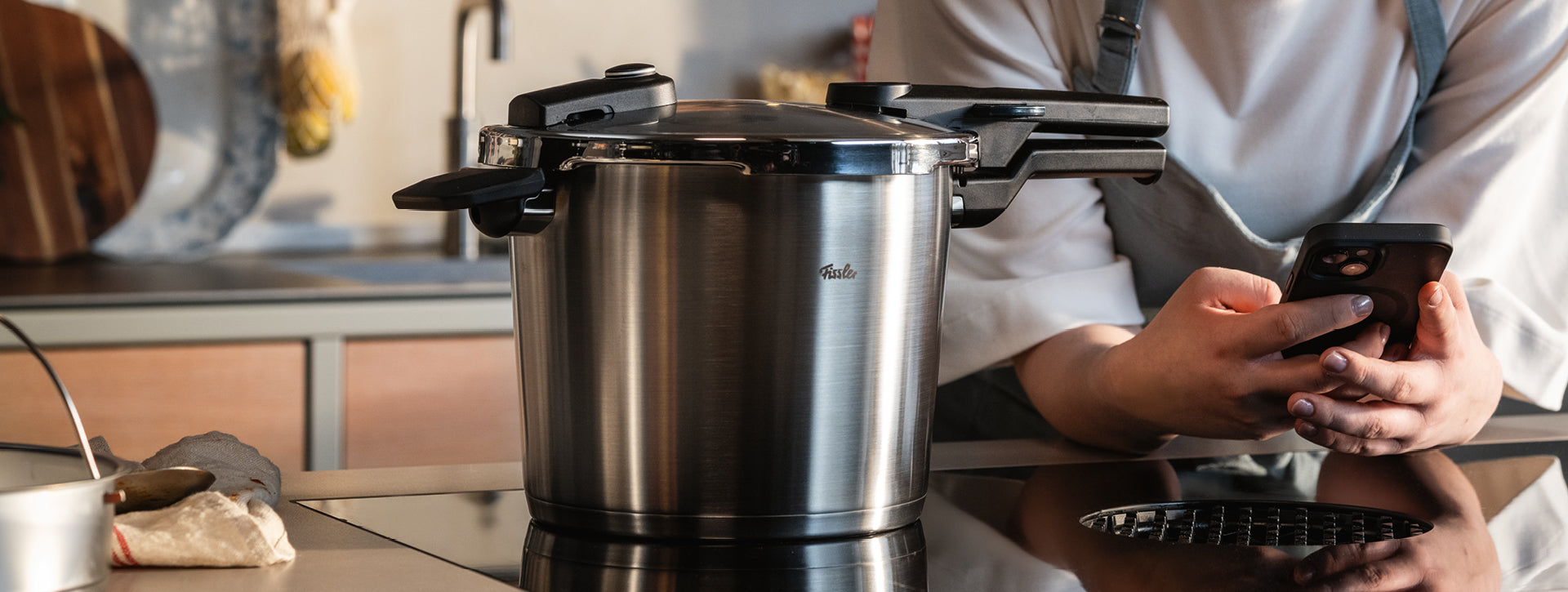 Eine Person in einer Küche benutzt ein Smartphone und lehnt sich neben einen Fissler Schnellkochtopf aus Edelstahl auf einer schlichten schwarzen Herdplatte.