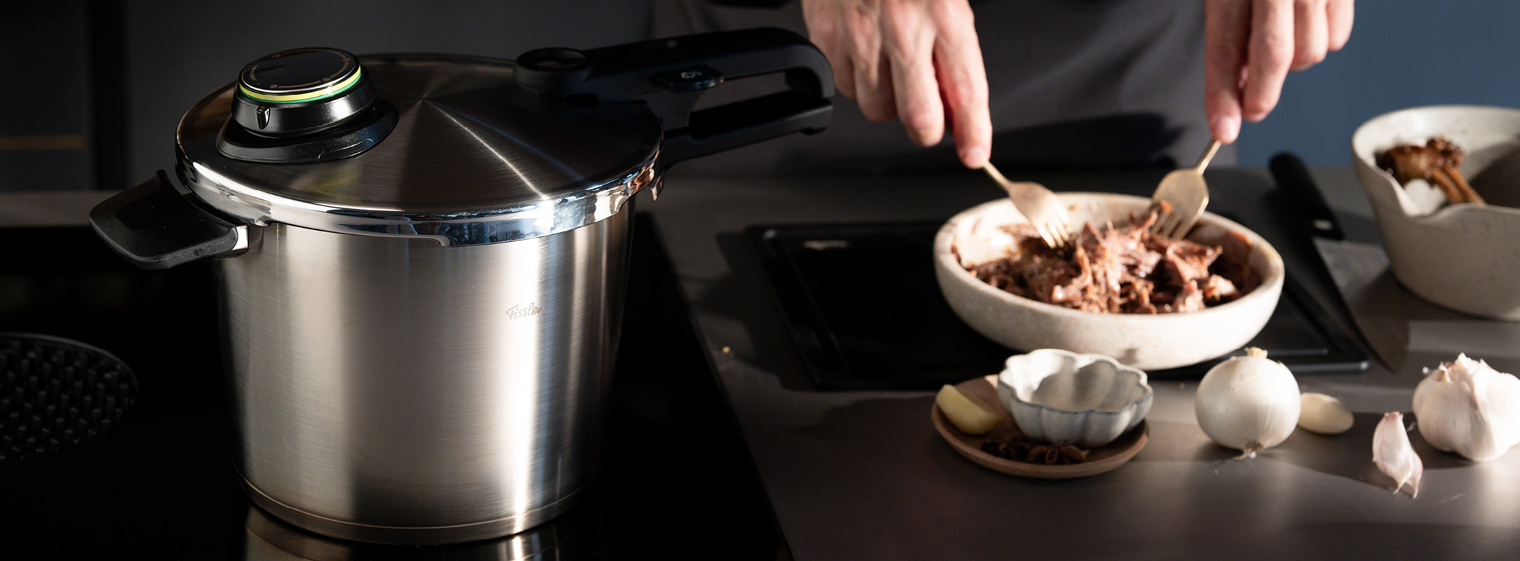 Ein Schnellkochtopf aus rostfreiem Stahl von Fissler auf einer Herdplatte, daneben wird Fleisch zerkleinert, umgeben von frischen Gewürzen und Zutaten.