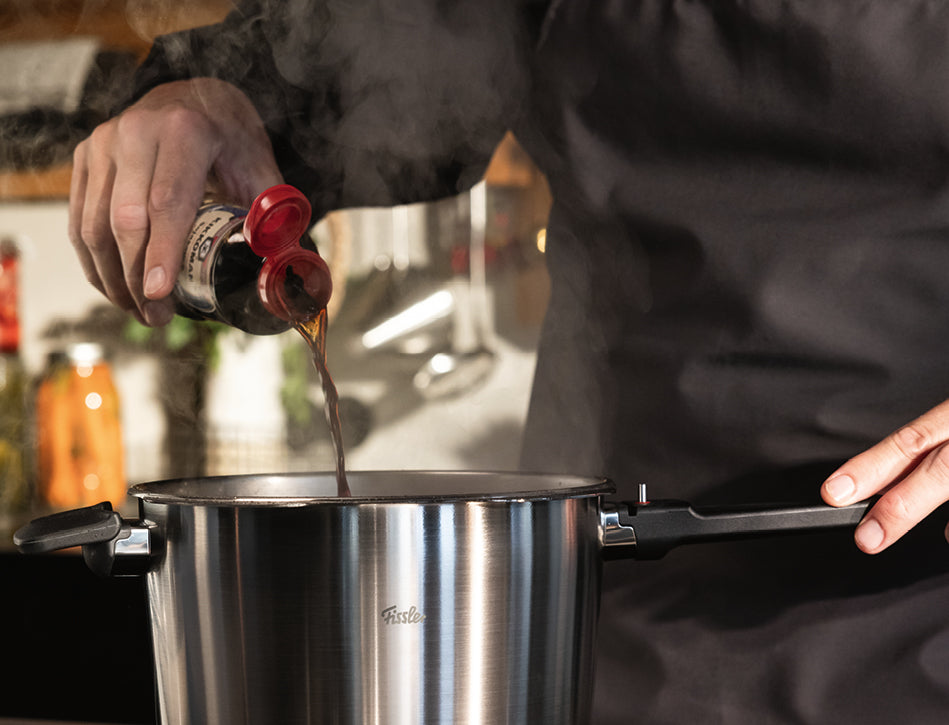 Eine Person in dunkler Schürze gibt Soße in einen dampfenden Fissler-Edelstahltopf und präsentiert deutsche Präzision und zeitloses Design am Herd.