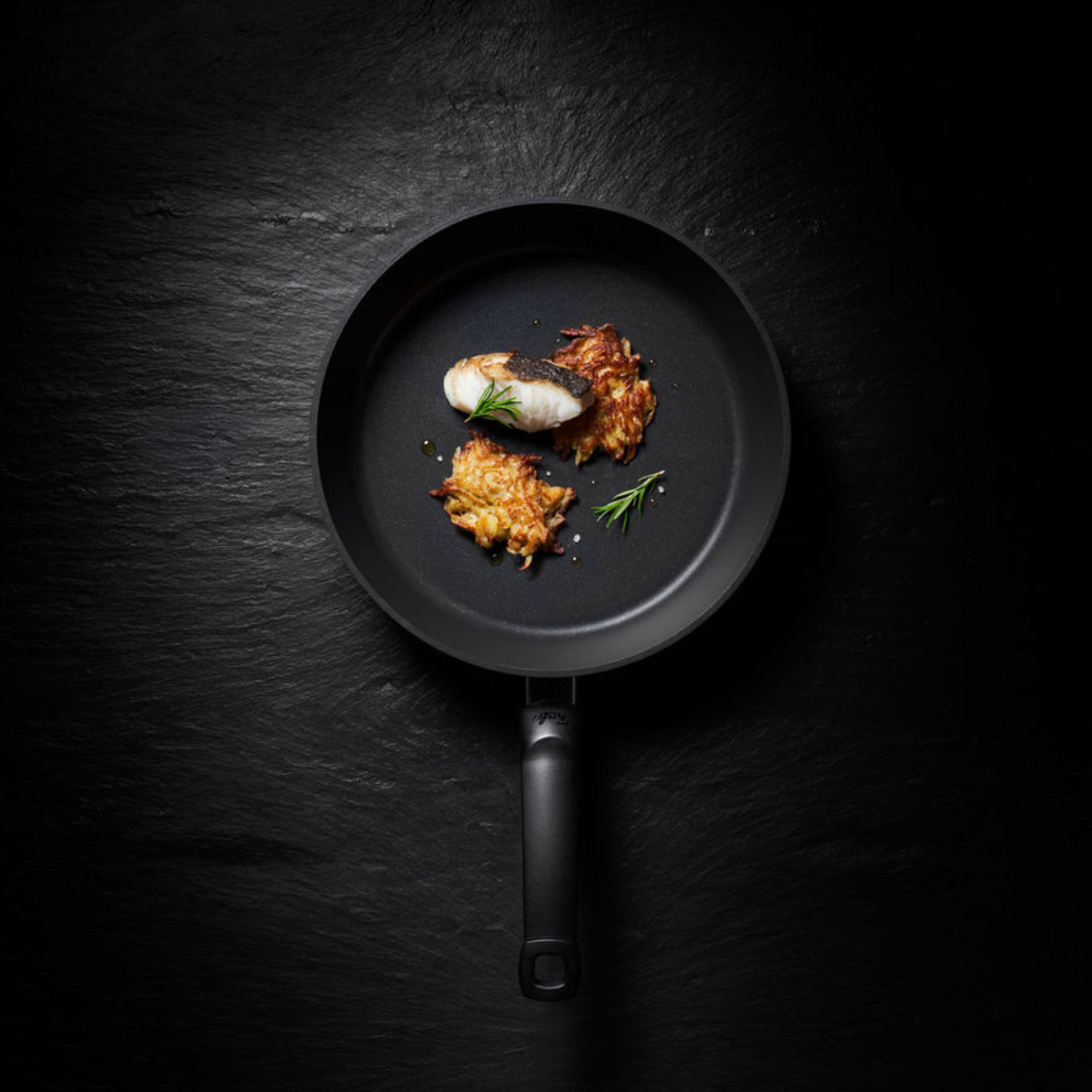 Eine Fissler Bratpfanne auf dunkler Oberfläche, in der Fisch, Kartoffelpuffer und Rosmarin elegant serviert werden - ein Beweis für deutsche Qualität und Design.