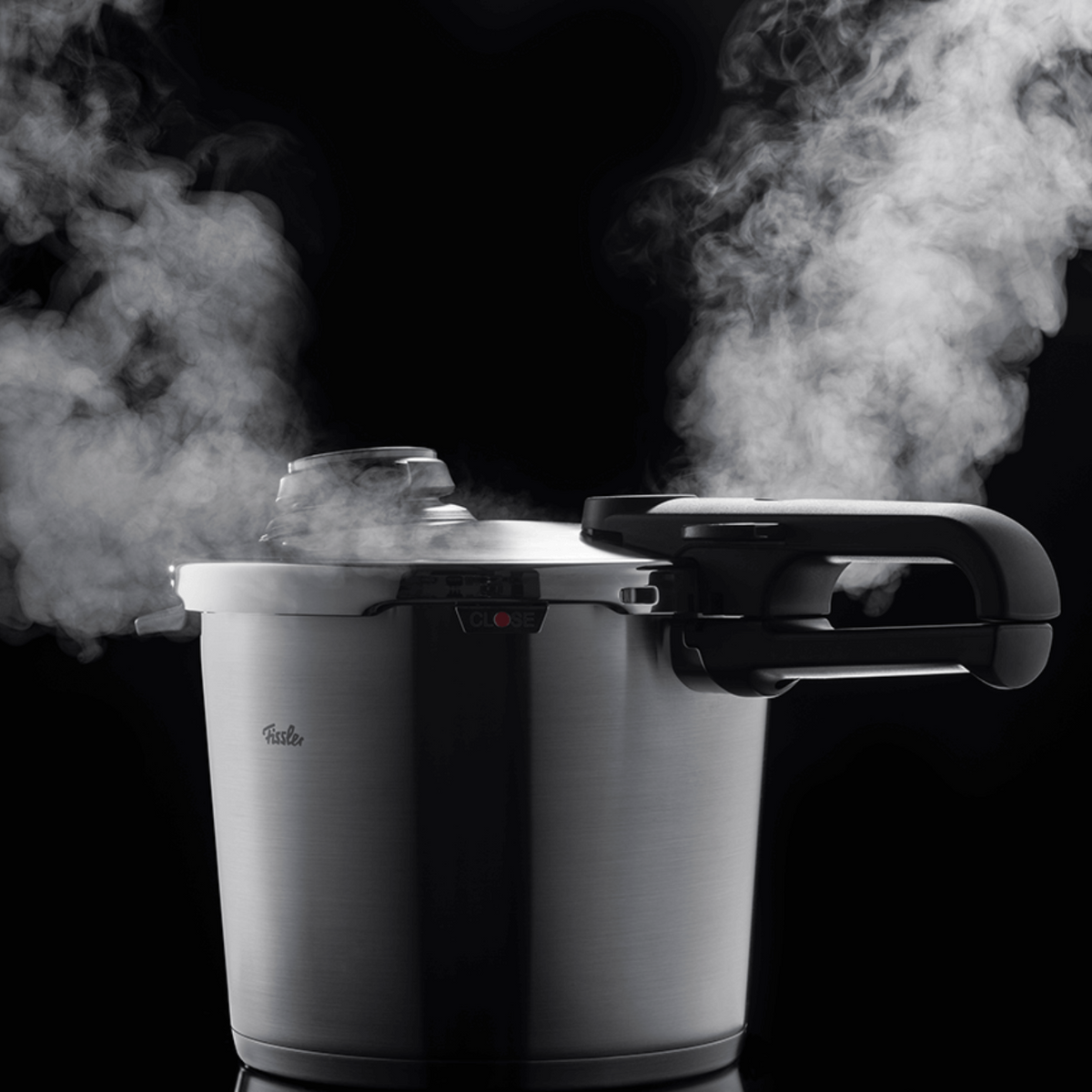 Ein Schnellkochtopf von Fissler aus rostfreiem Stahl mit schwarzen Griffen setzt Dampf frei und präsentiert deutsche Präzision und Qualität vor einem dunklen Hintergrund.