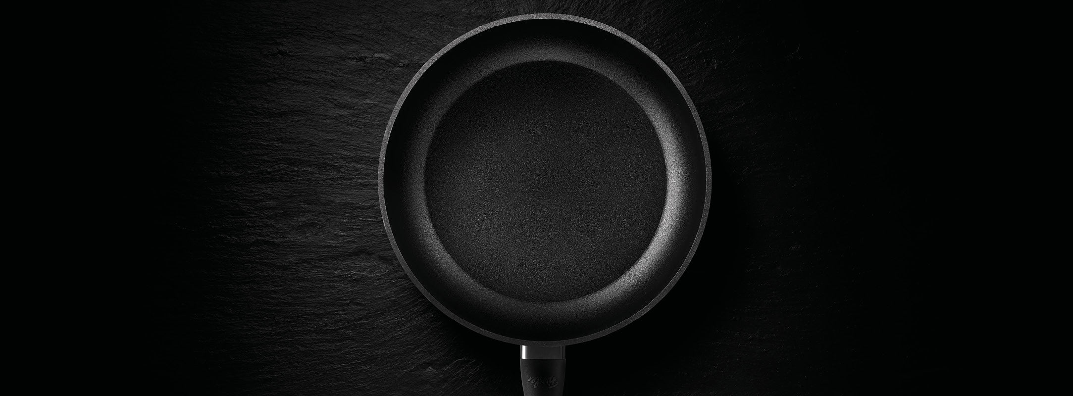 Eine schwarze antihaftbeschichtete Bratpfanne von Fissler in der Draufsicht, zentriert auf einem dunkel strukturierten Hintergrund - deutsche Präzisionsarbeit und zeitloses Design.