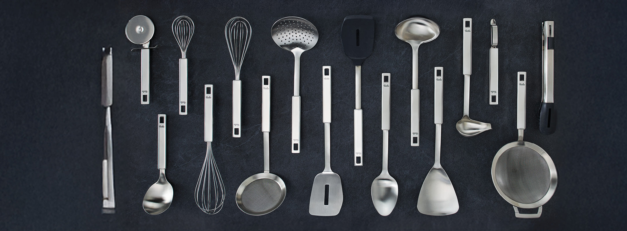Fissler Küchenutensilien aus rostfreiem Stahl, darunter Zangen, Schöpfkellen, Schneebesen und Pfannenwender, elegant angeordnet auf einem dunklen Hintergrund.
