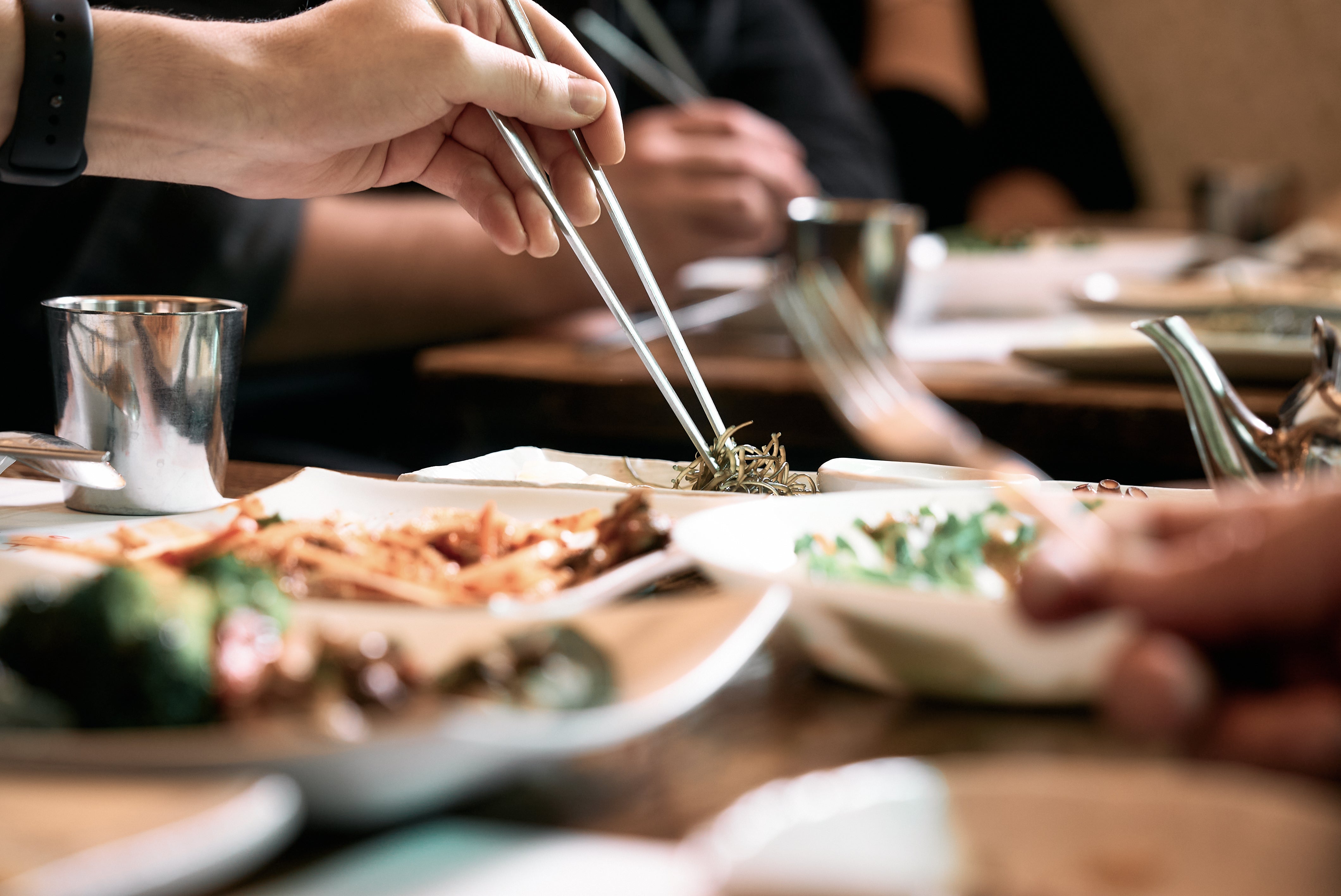 An einem wunderschön gedeckten Tisch, auf dem verschiedene Gerichte in elegantem Fissler-Edelstahlgeschirr serviert werden, nimmt eine Person mit Essstäbchen Essen vom Teller auf. Die Hände anderer greifen nach klassischen Fissler-Töpfen und -Pfannen – ein gemeinsames Mahl, das die präzise Technik, die Langlebigkeit und das zeitlose Design hochwertiger, in Deutschland hergestellter Küchenutensilien unterstreicht.