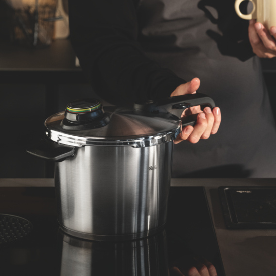 Eine Person hält einen Fissler-Schnellkochtopf auf der Herdplatte, eine andere Hand hält einen Becher. Der Küchenhintergrund ist sanft verschwommen.