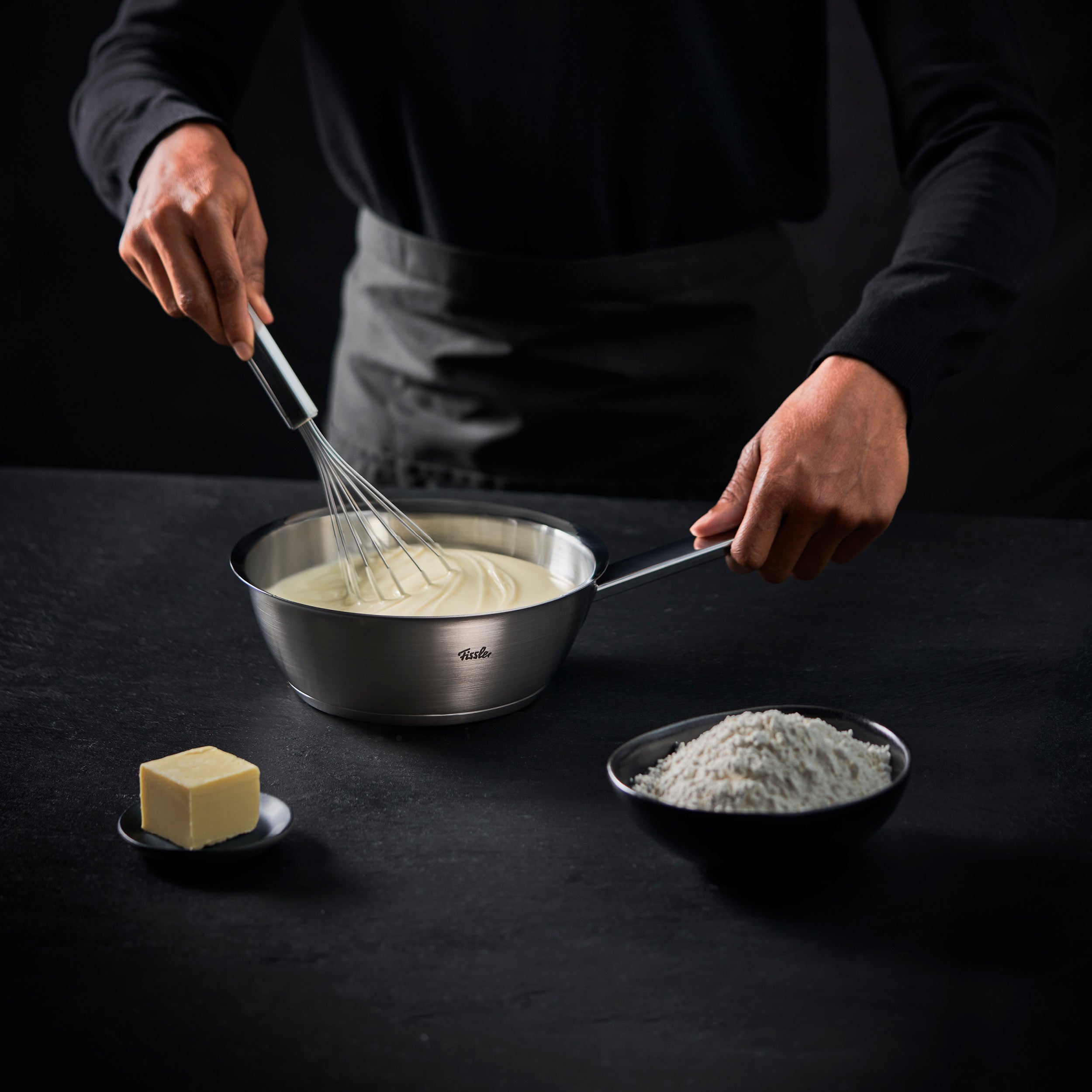 Mit einem Fissler-Schneebesen und einer Schüssel aus rostfreiem Stahl rührt ein Koch einen cremigen Teig. Butter und Mehl liegen in der Nähe, bereit für das Rezept.