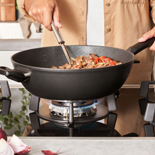 Eine Person kocht mit einem Fissler Adamant® Wok, bekannt für deutsche Präzision, auf einem Gasherd, während frische Zutaten brutzeln.