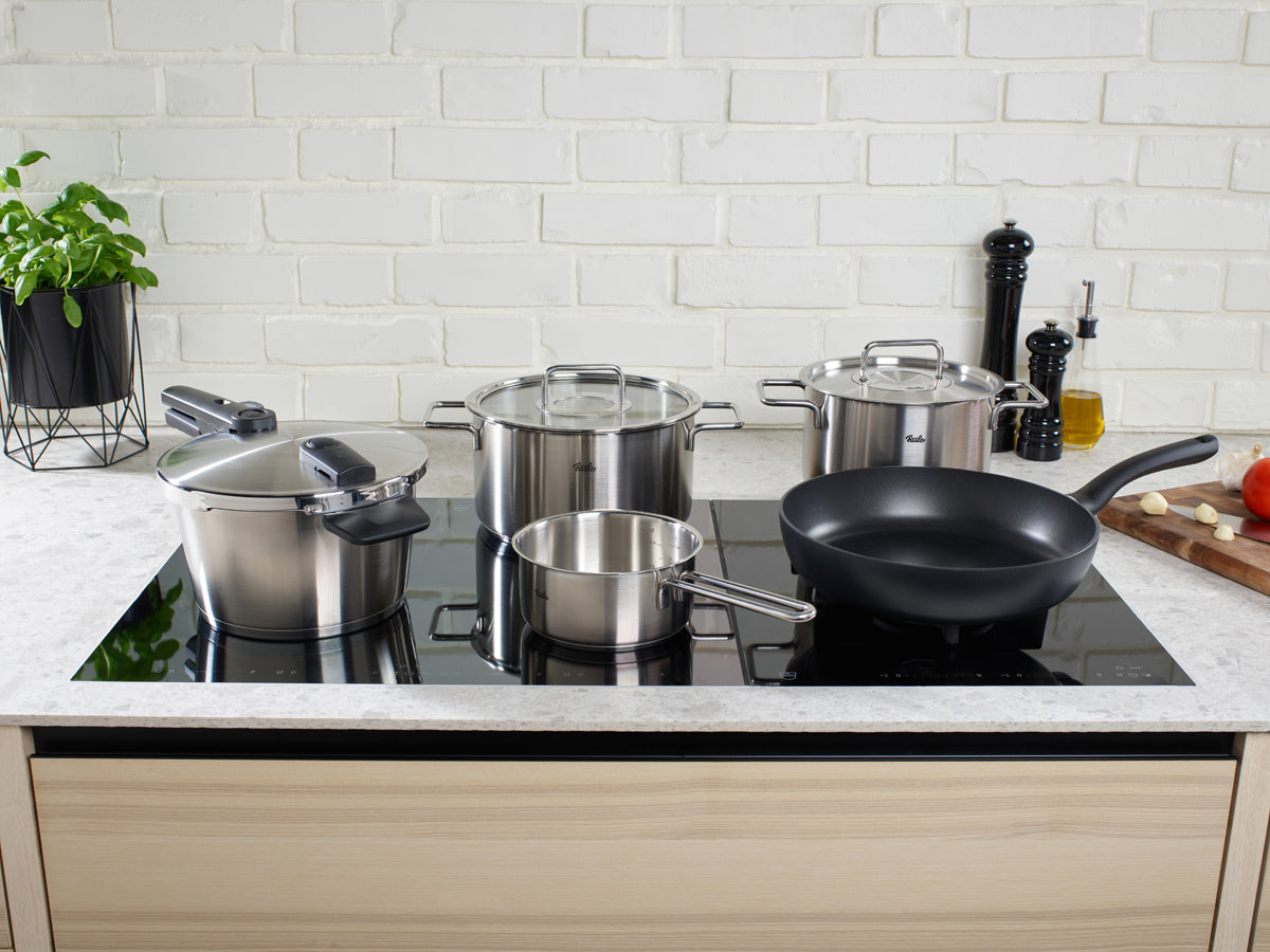 Fissler stainless steel pots and Cenit® Frying Pan on an induction hob, with fresh herbs and spices on a board beside them.