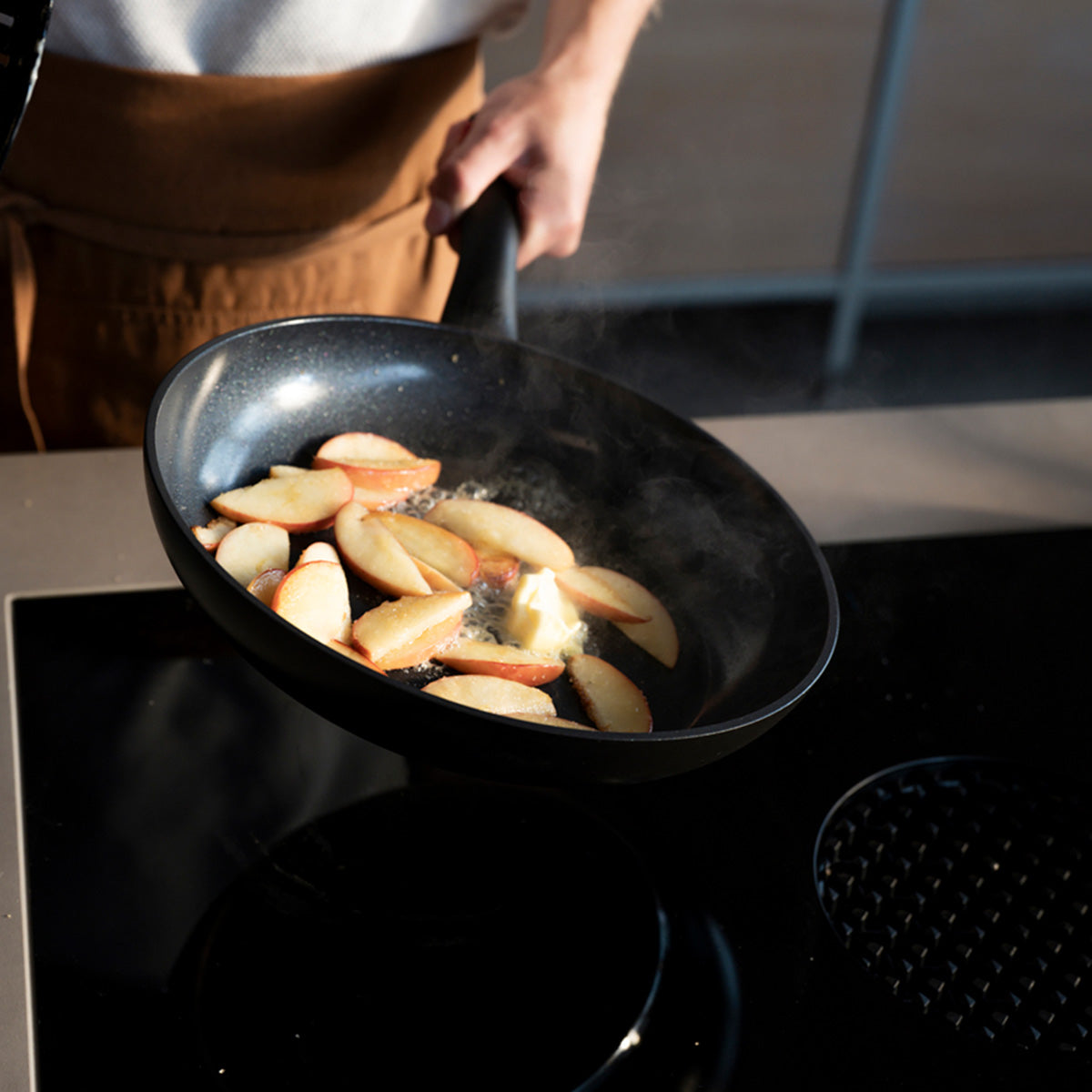 In Scheiben geschnittene Äpfel karamellisieren in einem Fissler Essential® Pfannenset, Dampf steigt auf, während Arm und Oberkörper des Kochs Präzision Made in Germany" zeigen.