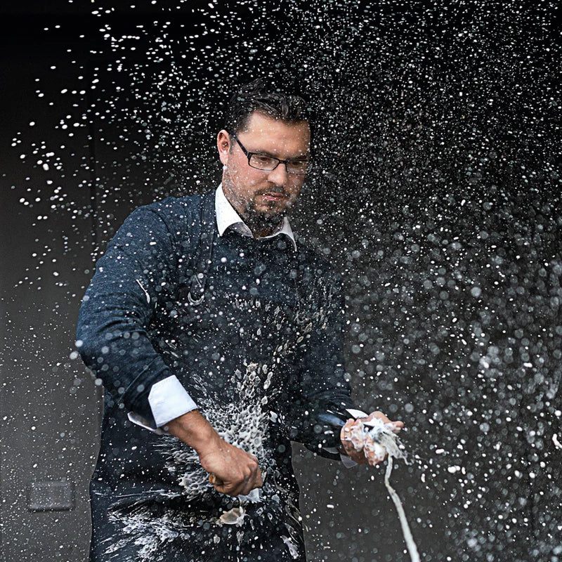 Ein Mann mit Brille und Kochschürze demonstriert präzise Kochtechnik, während er einen weißen Gegenstand zusammendrückt, woraufhin ein beeindruckender Sprühnebel aus Flüssigkeitstropfen entsteht. Diese Szene unterstreicht Fisslers Engagement für hochwertiges Kochgeschirr und innovatives Design und spiegelt die hervorragende Handhabung und Leistung wider, die unsere Küchenutensilien aus Edelstahl bieten.