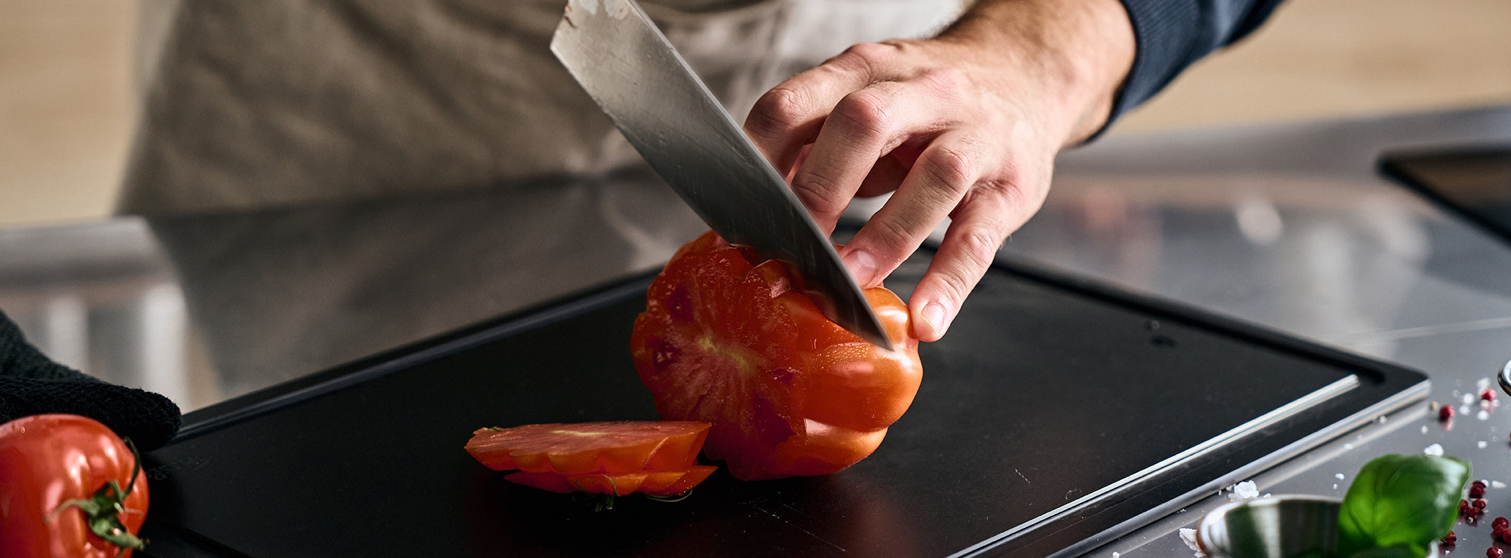 Eine Person schneidet eine Tomate auf einem Fissler-Schneidebrett präzise in Scheiben, während in der Nähe Fissler-Messer, frische Kräuter und Küchengeräte ausgestellt sind.