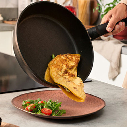 A hand tilts the Fissler Levital®+ Comfort Pan and serves an omelet on a plate - precision, durability, and quality Made in Germany.