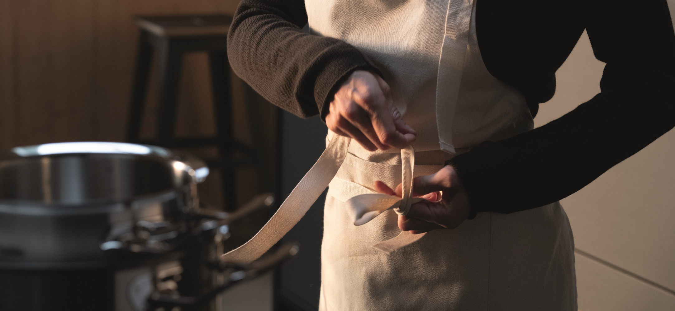 Eine Person bindet eine beigefarbene Schürze in einer Küche, im Hintergrund sind ein hochwertiger Fissler-Edelstahltopf und ein Hocker zu sehen.