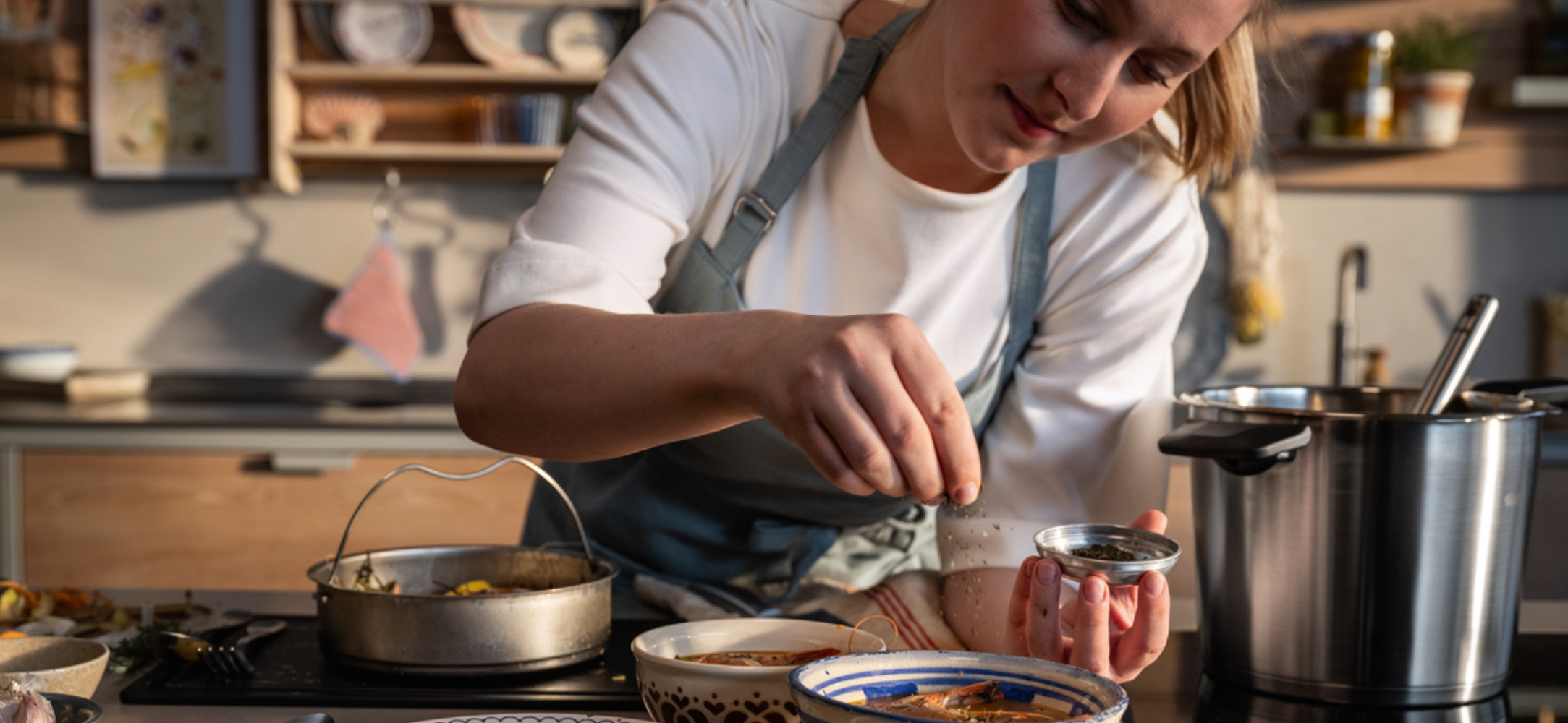 Eine Person in einer Schürze bereitet das Essen zu und streut Kräuter in eine Schüssel, die von Fissler Kochgeschirr und Küchenzubehör umgeben ist.