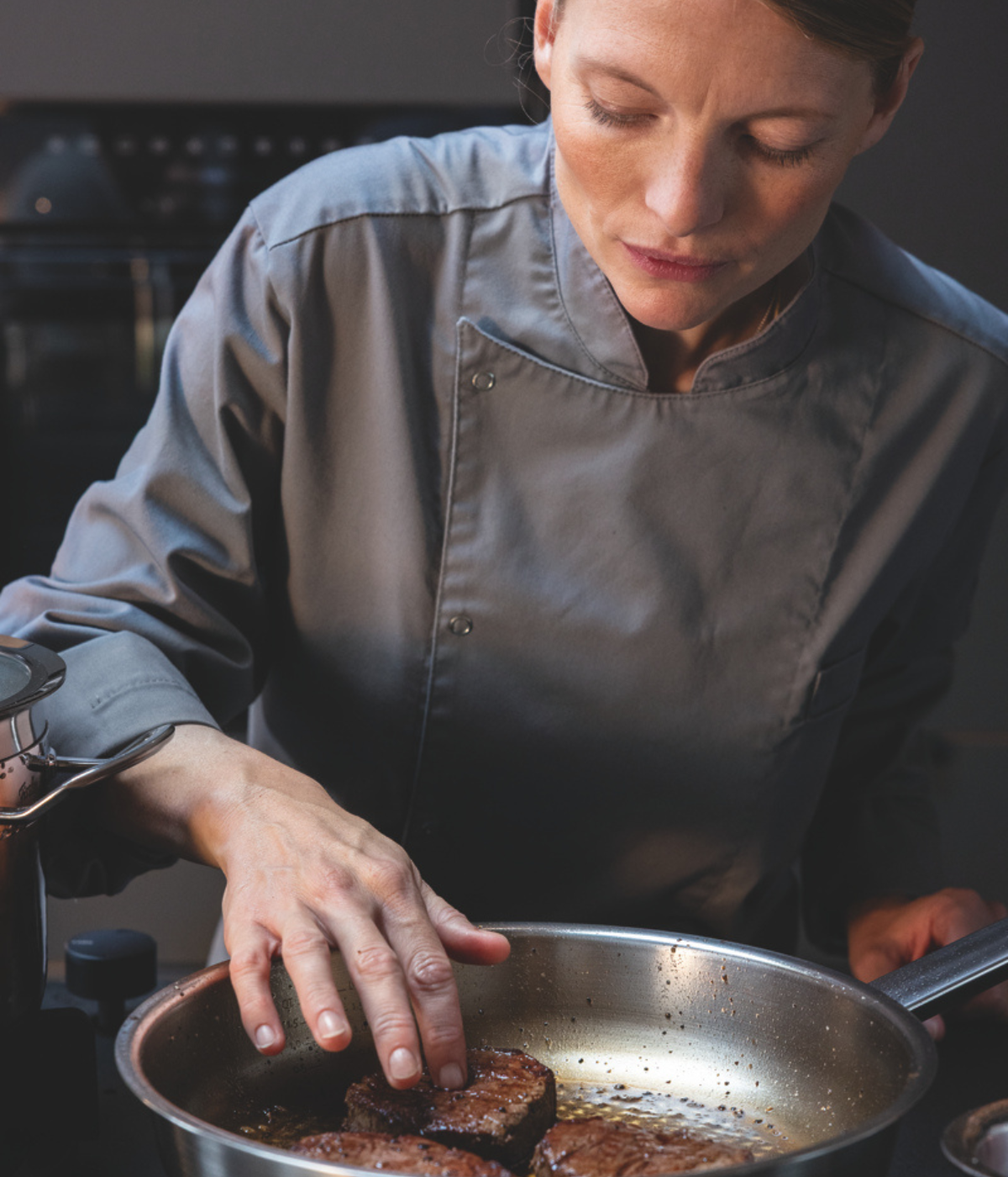 A chef in a gray uniform carefully checks the doneness of steak sizzling in a premium Fissler stainless steel frying pan, highlighting the precision and quality of Fissler’s German-engineered cookware.