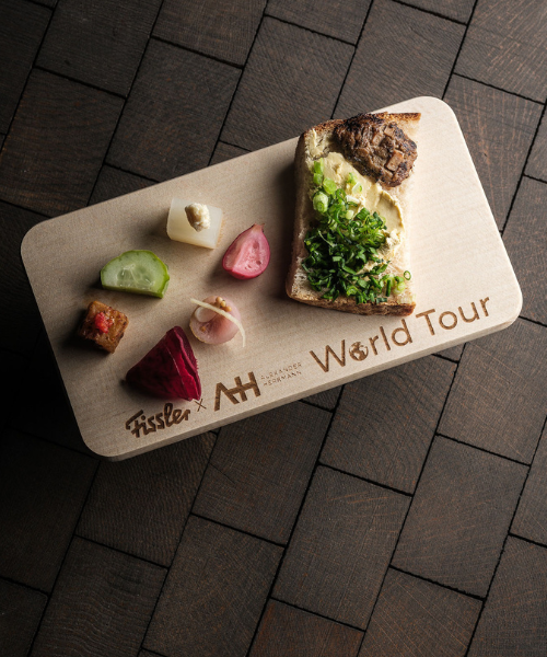 Auf einem präzise gefertigten Holzbrett, das mit der Aufschrift „Fissler x AH World Tour“ graviert ist, werden elegant eine Scheibe Brot mit frischen Frühlingszwiebeln sowie fünf kunstvoll angerichtete, farbenfrohe Häppchen präsentiert – alles auf einer eleganten, dunklen Fliesenoberfläche. Die raffinierte Präsentation spiegelt Fisslers Bekenntnis zu zeitlosem Design und deutscher Ingenieurskunst in der Herstellung hochwertiger Kochgeschirr-Handwerkskunst wider.