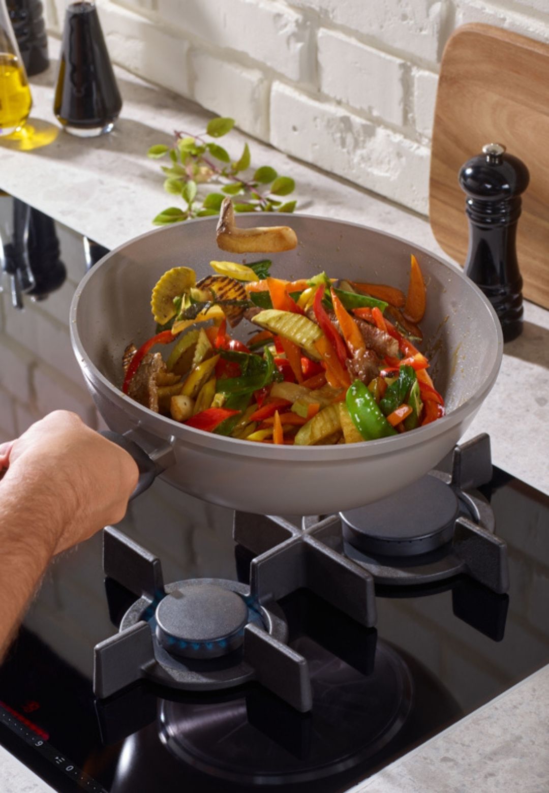 Eine Person kocht lebhaftes Gemüse in einer Fissler-Pfanne auf einem Gasherd, eine moderne Küche mit weißen Backsteinwänden und Utensilien dahinter.