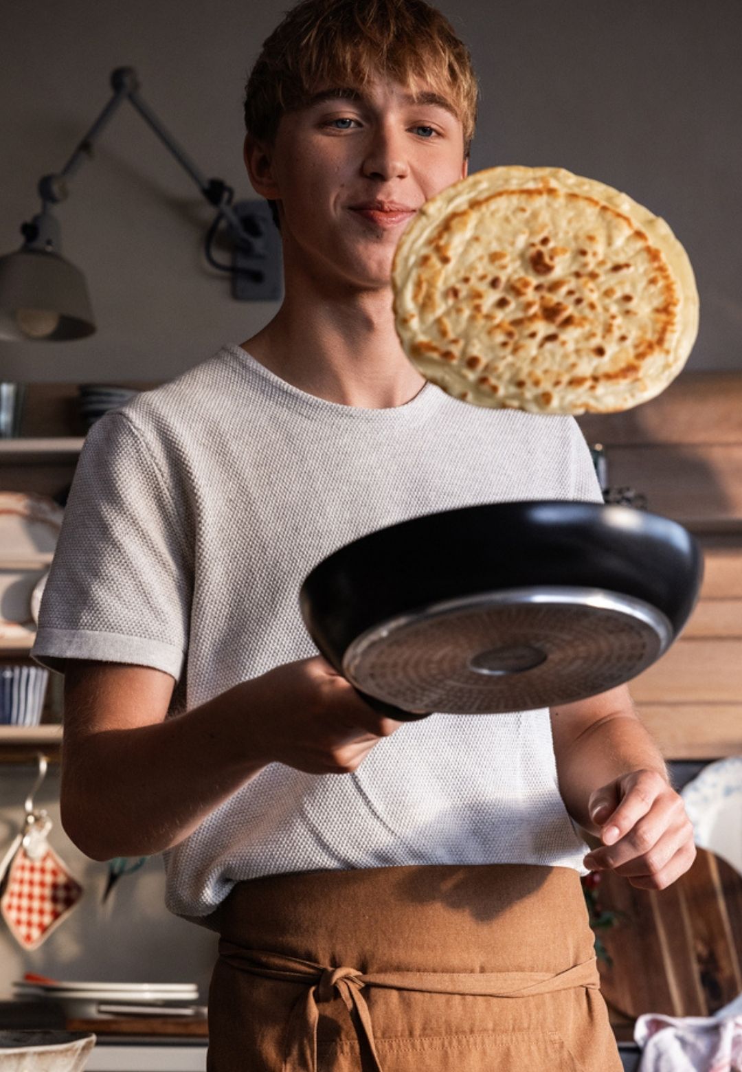 Eine junge Person wendet einen Pfannkuchen in einer schwarzen Bratpfanne von Fissler, die deutsche Qualität und zeitloses Design in einer gemütlichen Küche präsentiert.
