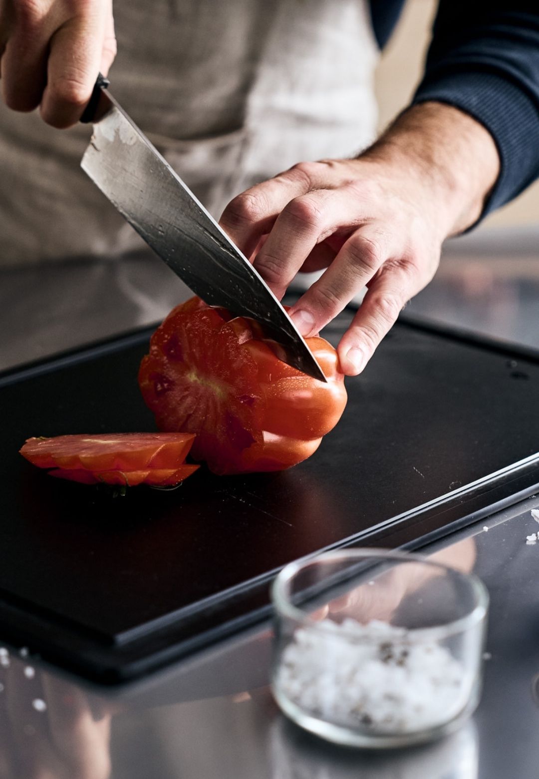 Ein Koch schneidet eine Tomate mit Präzision auf einem Fissler-Schneidebrett in Scheiben, während auf der glatten Arbeitsplatte grobes Salz in einer Glasschale liegt.