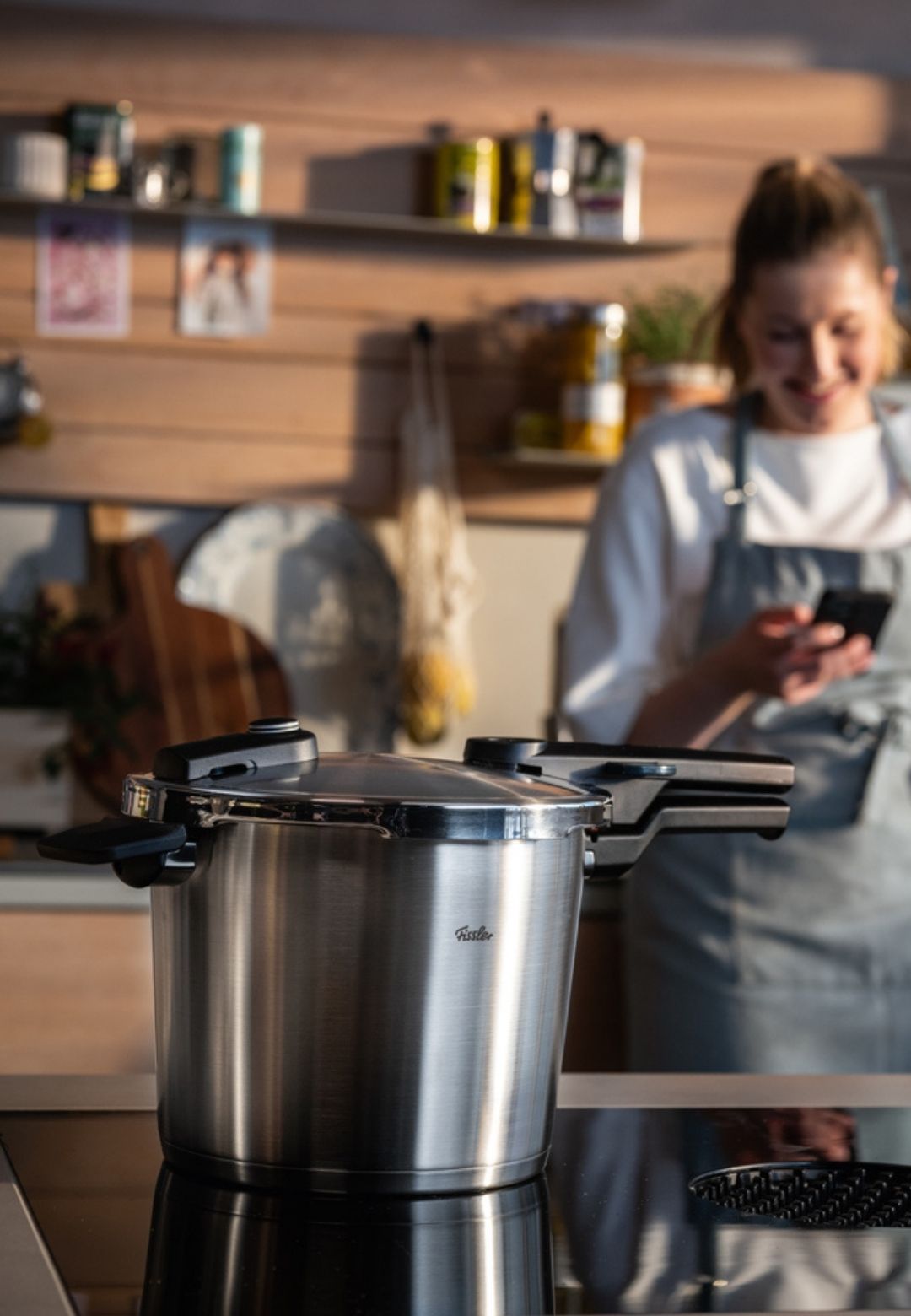 Ein Fissler Schnellkochtopf aus Edelstahl auf einer modernen Herdplatte; eine Frau lächelt in der Nähe und hebt deutsche Qualität und Innovation hervor.