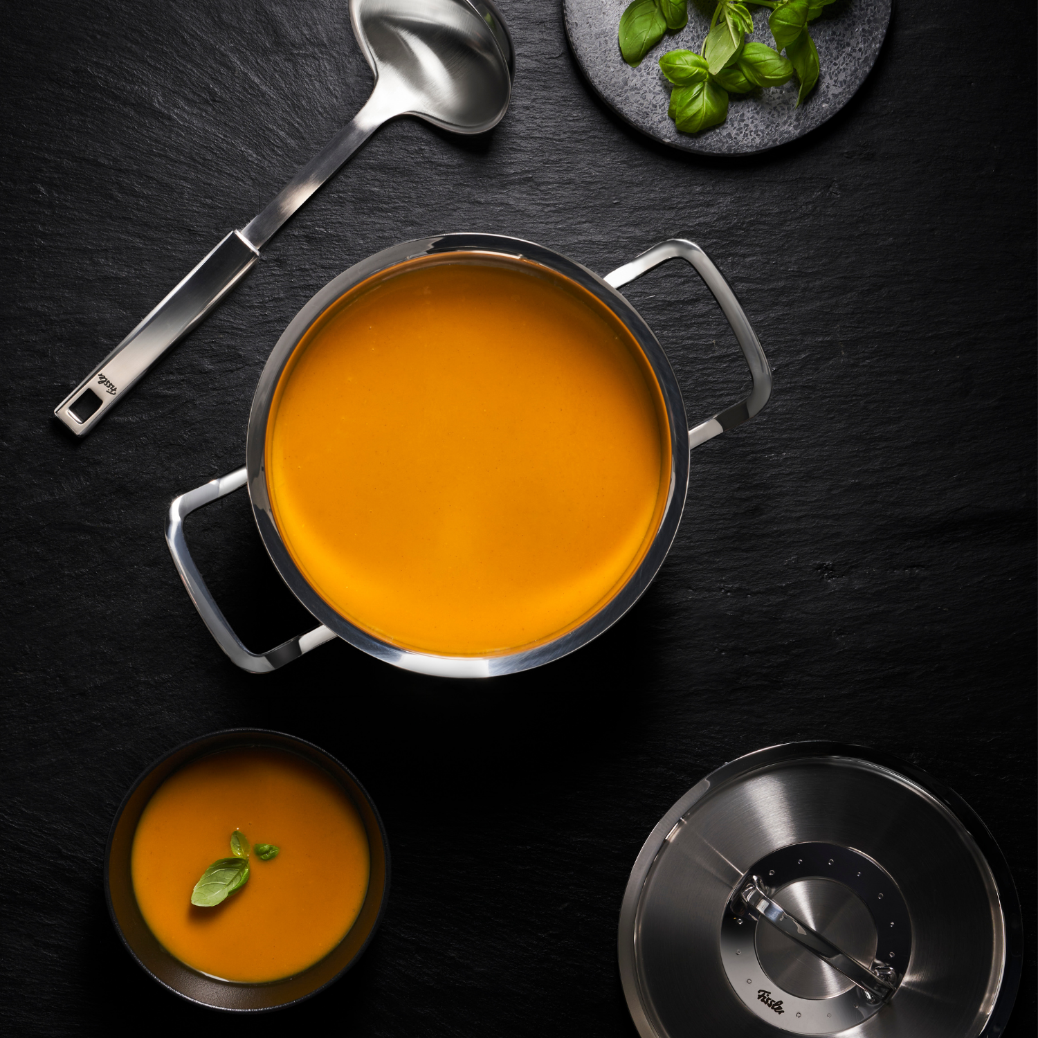 Fissler-Edelstahltopf mit cremiger Suppe auf einer dunklen Fläche, Deckel und Schöpfkelle daneben; Schale mit Suppe, garniert mit Basilikum.