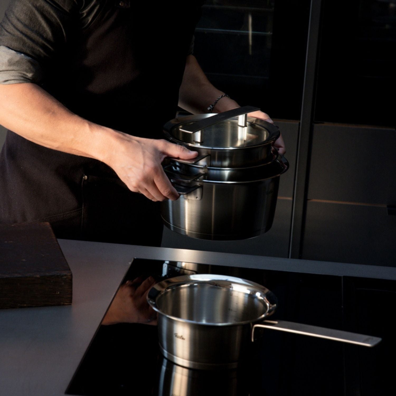 Eine Person hält zwei gestapelte Fissler-Edelstahltöpfe in einer modernen Küche; ein weiterer Fissler-Topf steht auf einem schwarzen Induktionskochfeld.