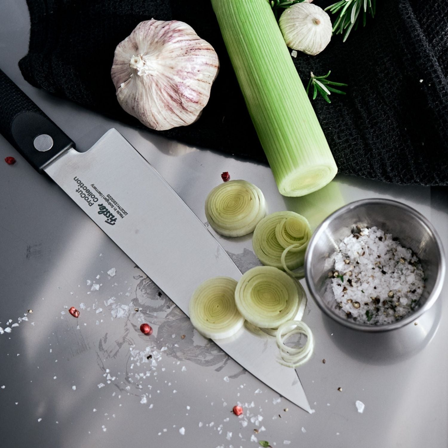 Ein präzises Fissler-Kochmesser aus rostfreiem Stahl mit frischem Lauch, Knoblauch, Rosmarin und einer Stahltasse mit grobem Salz und Pfeffer.