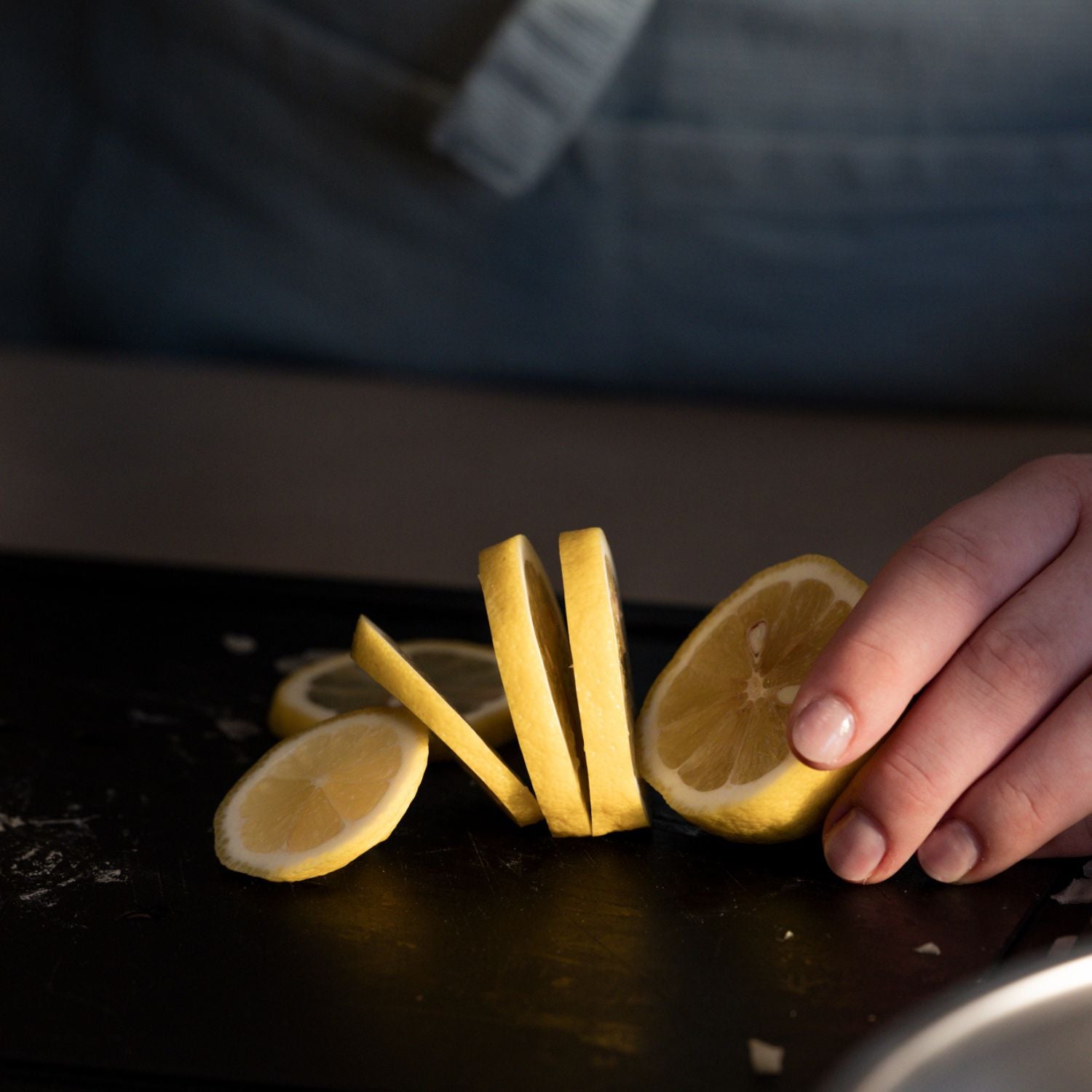 Eine Hand bereitet eine Zitrone auf einem glatten schwarzen Fissler-Schneidebrett zu, wobei die Scheiben kunstvoll angeordnet sind; die Person trägt eine blaue Schürze.