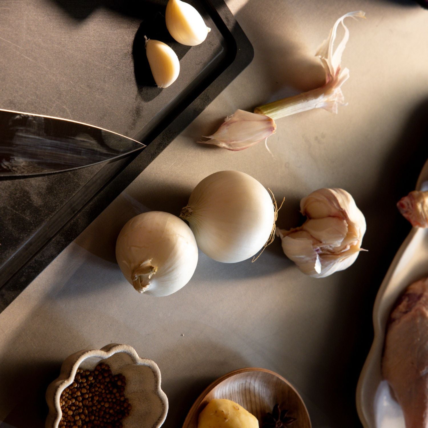 Blick von oben auf eine Küchentheke mit frischen Zwiebeln, Knoblauch, Gewürzen und rohem Fleisch neben hochwertigem Fissler-Geschirr.
